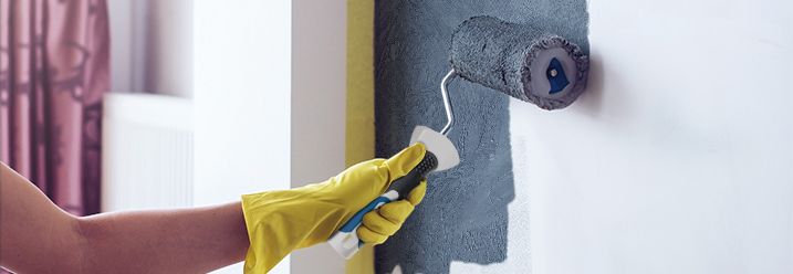 Eine Frau mit gelbem Malerhandschuh streicht eine Wand in grau.
